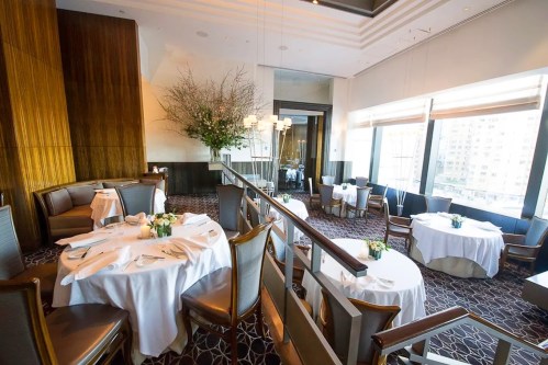 Elegant restaurant interior with round tables, white tablecloths, and large windows offering a city view.
