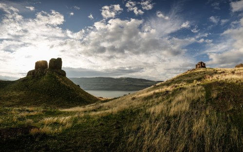 A scenic landscape featuring grassy hills, rocky formations, and a cloudy sky over a calm body of water.