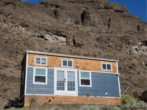A small, modern house with a wooden exterior sits at the base of a rocky mountain landscape.