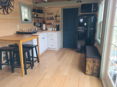 Cozy kitchen with wooden floors, white cabinets, black fridge, and a small dining table with stools. Natural light from windows.