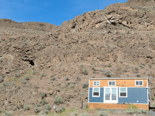 A small blue and wood-paneled house sits at the base of a rocky, mountainous landscape under a clear blue sky.