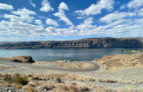 A winding road curves along a rocky shoreline with a calm lake and cliffs under a blue sky with scattered clouds.