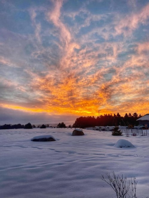 A vibrant sunset casts orange and pink hues over a snowy landscape, with rocks and trees silhouetted against the sky.