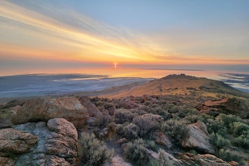 A serene sunset over a vast landscape, featuring rocky terrain and a shimmering body of water in the distance.