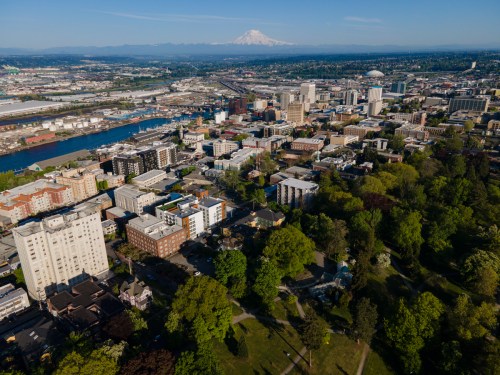 Aerial view of a city with buildings, trees, and a river, with mountains in the background under a clear blue sky.