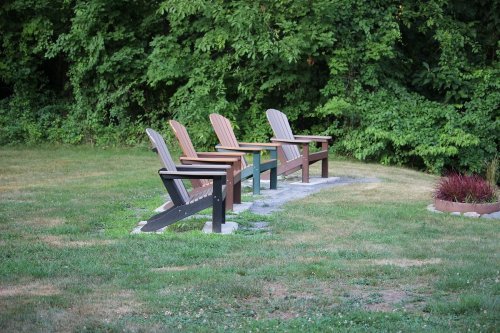 Four wooden Adirondack chairs in a grassy area, set against a backdrop of lush green trees.