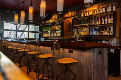 A stylish bar with wooden stools, shelves lined with various bottles, and modern pendant lights hanging above.