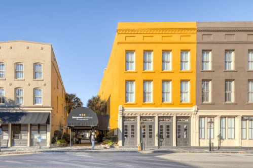 A vibrant yellow building stands next to a brown building, with a canopy entrance and palm trees in the background.