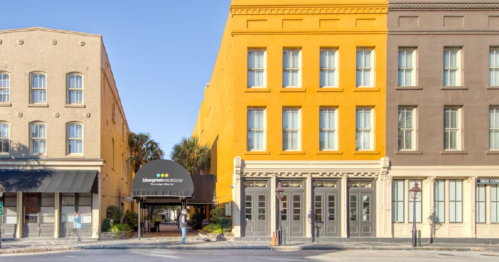 A vibrant yellow building stands between two other buildings, with a canopy entrance and palm trees in the background.