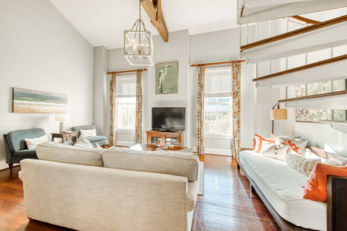 Bright living room with a high ceiling, large windows, a TV, and a staircase, featuring cozy furniture and decorative accents.