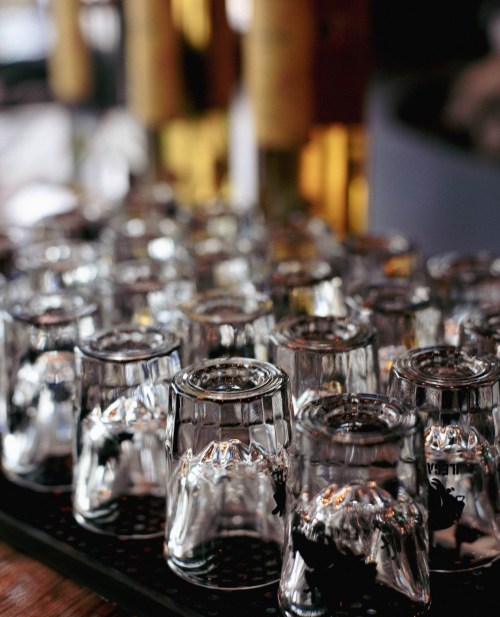 A close-up of a tray filled with clear glass shot glasses, with blurred bottles in the background.