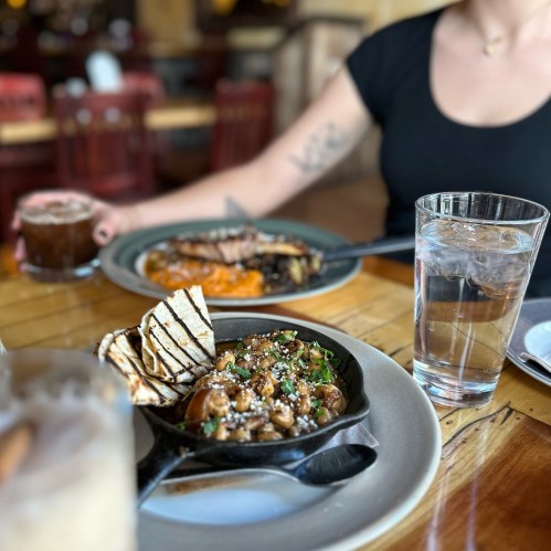 A table setting with a cast iron skillet of food, grilled bread, a glass of water, and a person in a black shirt.
