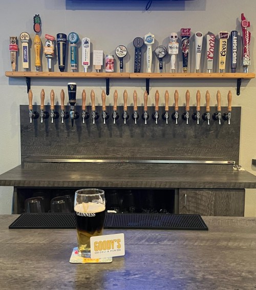 A bar with a variety of beer taps and a glass of Guinness on the counter, featuring a card that says "GOODY'S."