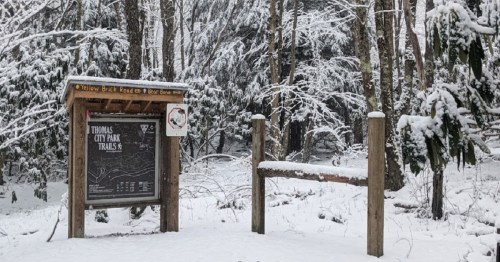 Snow-covered sign for Thomas City Park Trails, surrounded by trees in a winter landscape.