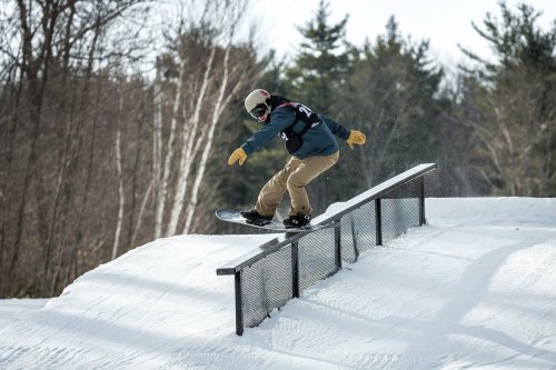 A snowboarder performs a trick on a rail in a snowy terrain, surrounded by trees.