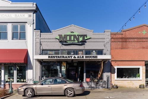 Exterior of The Mint Restaurant and Ale House, featuring a gray facade and outdoor seating, with a clear blue sky above.