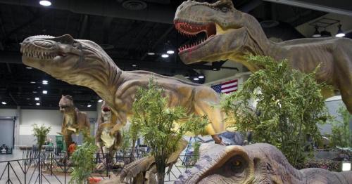 Three large dinosaur models displayed indoors among greenery, with an American flag visible in the background.