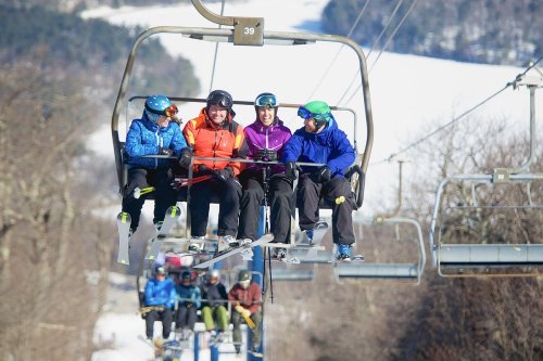 A group of four skiers on a chairlift, wearing colorful ski gear, enjoying a winter day in the mountains.