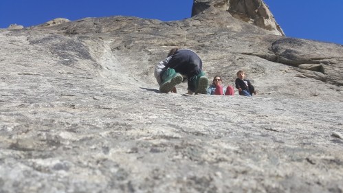 A climber ascends a rocky surface while two people sit on the ledge above, enjoying the sunny day.
