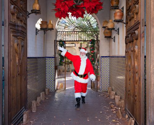 Santa Claus in a red suit waves cheerfully while walking through a festive archway decorated with poinsettias.