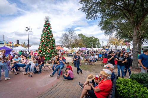 A festive outdoor event with a large Christmas tree, crowds of people, and colorful tents under a cloudy sky.