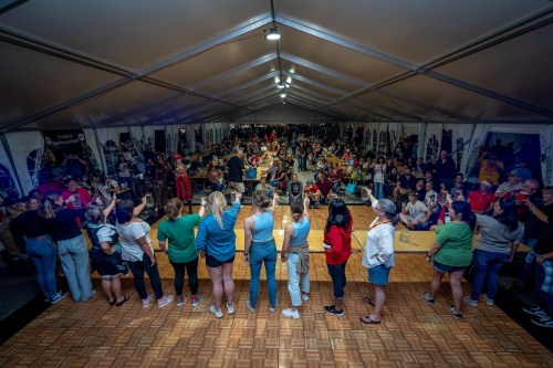 A large crowd gathers under a tent, with people facing the audience, enjoying an event or celebration.