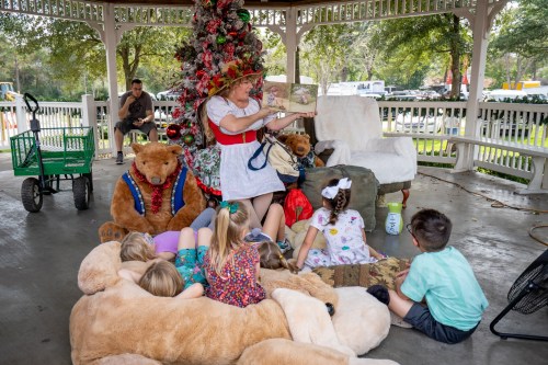 A storyteller in a festive hat reads to children sitting on a large teddy bear and a cozy rug under a decorated gazebo.