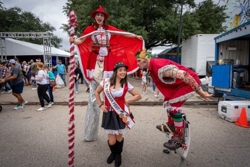 A festival scene with a woman in a sash posing with stilt walkers in costumes, surrounded by a crowd and festive decorations.