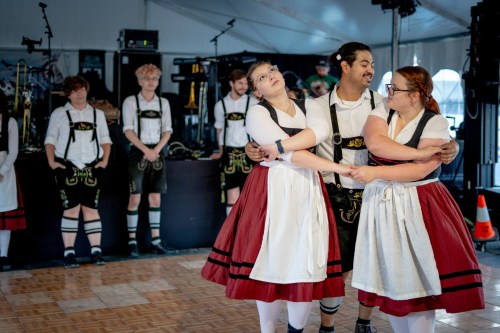 Three dancers in traditional attire perform in a tent, while a band and other performers watch in the background.