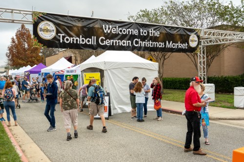 A bustling outdoor market scene with a welcome banner for the Tomball German Christmas Market and various tents.
