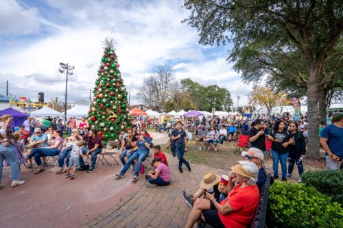 A festive outdoor event with a large Christmas tree, crowds of people, and colorful tents under a cloudy sky.