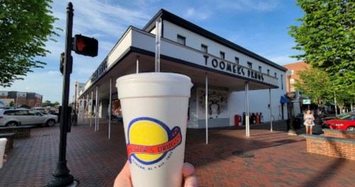 A hand holds a drink cup in front of Toomer's Drugs, a historic building with a blue sky and trees nearby.