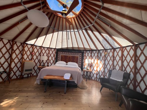 Cozy yurt interior with a bed, wooden furniture, and large windows letting in natural light.