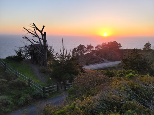 Sunset over the ocean, with a silhouette of trees and a winding road in the foreground.