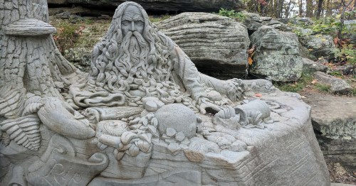A detailed stone sculpture of a bearded figure surrounded by nature, with intricate carvings of plants and animals.