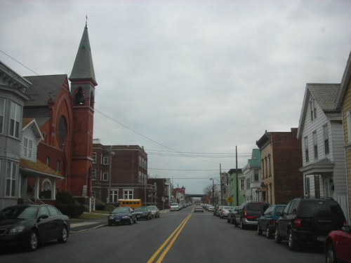A street view featuring a red church, lined with houses and parked cars under a cloudy sky.