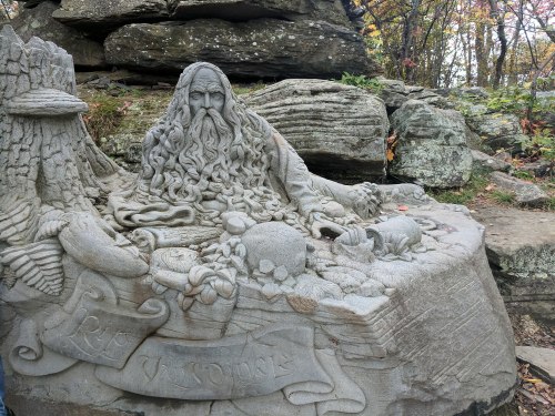A detailed stone carving of a bearded figure surrounded by nature, fruits, and animals, set among rocky terrain.