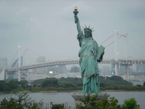 The Statue of Liberty stands tall with a bridge and city skyline in the background, under a cloudy sky.