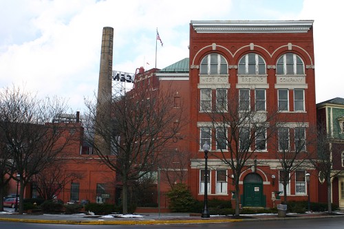Historic red brick building with "West End Brewing Company" sign, featuring a tall smokestack and trees in front.