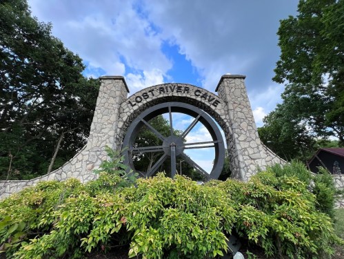 Stone entrance to Lost River Cave featuring a large wheel and surrounded by greenery under a cloudy sky.