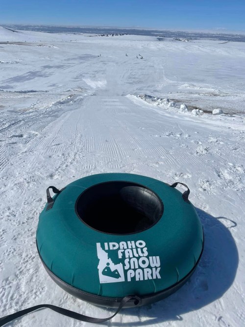 A green snow tube sits at the top of a snowy slope at Idaho Falls Snow Park, with distant figures enjoying the hill.