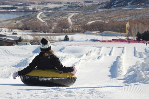 A person in a black jacket and hat sits on a snow tube, ready to slide down a snowy hill with a scenic background.