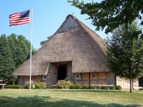 A thatched-roof barn with a flagpole displaying the American flag, surrounded by greenery and a well-maintained lawn.