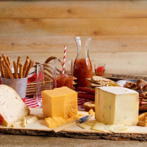 A wooden platter with various cheeses, pretzel sticks, drinks, and snacks arranged on a rustic table.