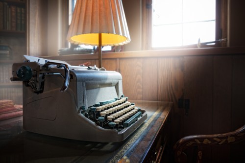 A vintage typewriter sits on a desk beside a lamp, with warm light illuminating a cozy room.
