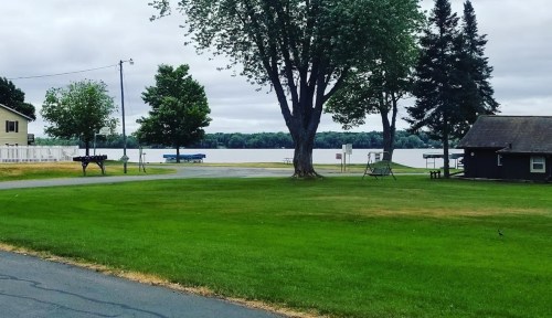 A serene lakeside view with green grass, trees, and a cloudy sky, featuring a small dock and a few boats.