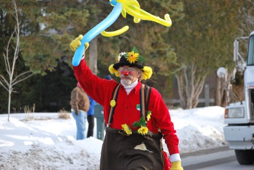 A cheerful clown in a red shirt and yellow gloves waves a blue balloon in a snowy outdoor setting.