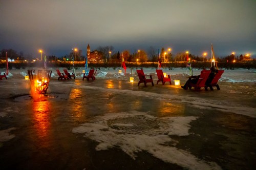 Red chairs lined up by a frozen lake, with lanterns and a fire pit glowing against a twilight sky.