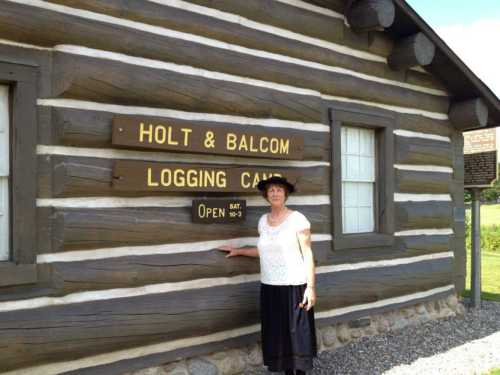 A woman in a hat stands by a wooden building with signs for "Holt & Balcom Logging Camp" and "Open Sat 10-3."