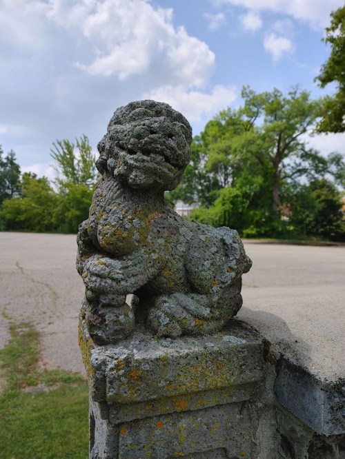 A weathered stone lion statue sits on a stone pedestal, surrounded by greenery and a cloudy sky.